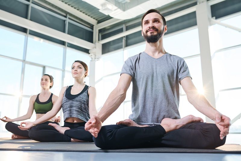 3 pratiquants du yoga, un homme en avant plan, assis en tailleur